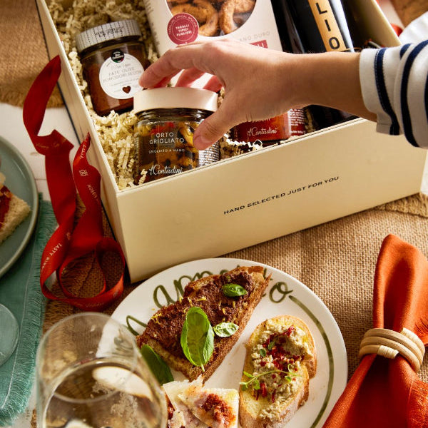 A gourmet gift box set displayed on a rustic burlap tablecloth, featuring a cream-colored box filled with assorted treats including a jar of prosciutto crudo, a bottle of Orgolio balsamic vinegar, and a box of Le Contadini focaccia. A hand is shown reaching for a jar lid, with plates of appetizers and a glass of white wine in the foreground.